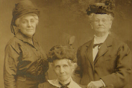 Portrait of three women wearing nice clothes and hats, one of whom is sitting