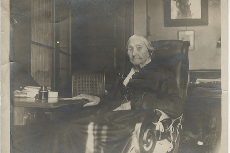 Photograph of a person sitting at a table with papers and an inkwell on it and their room in the background