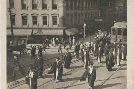 Aged photograph of a parade of women in academic caps and gowns marching along a raised roadway in front of a streetcar. A busy street and a billiard academy are visible in the background.